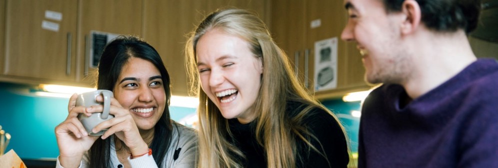 A photograph of a diverse group of students laughing together as they drink coffee in the kitchen of their halls of residence.